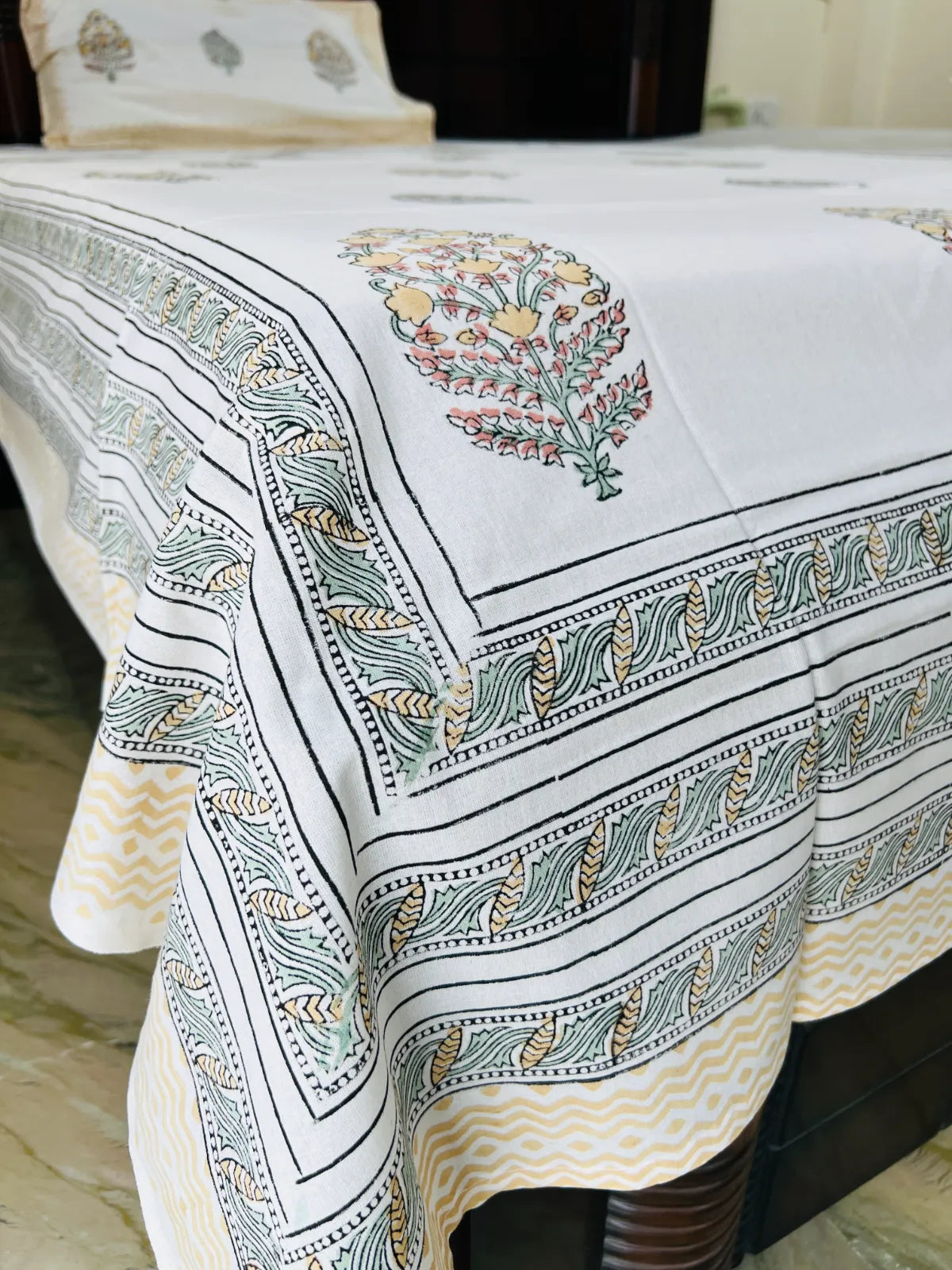 A white cotton bedspread with traditional block-printed floral and geometric patterns in black, green, and yellow, draped over a bed.