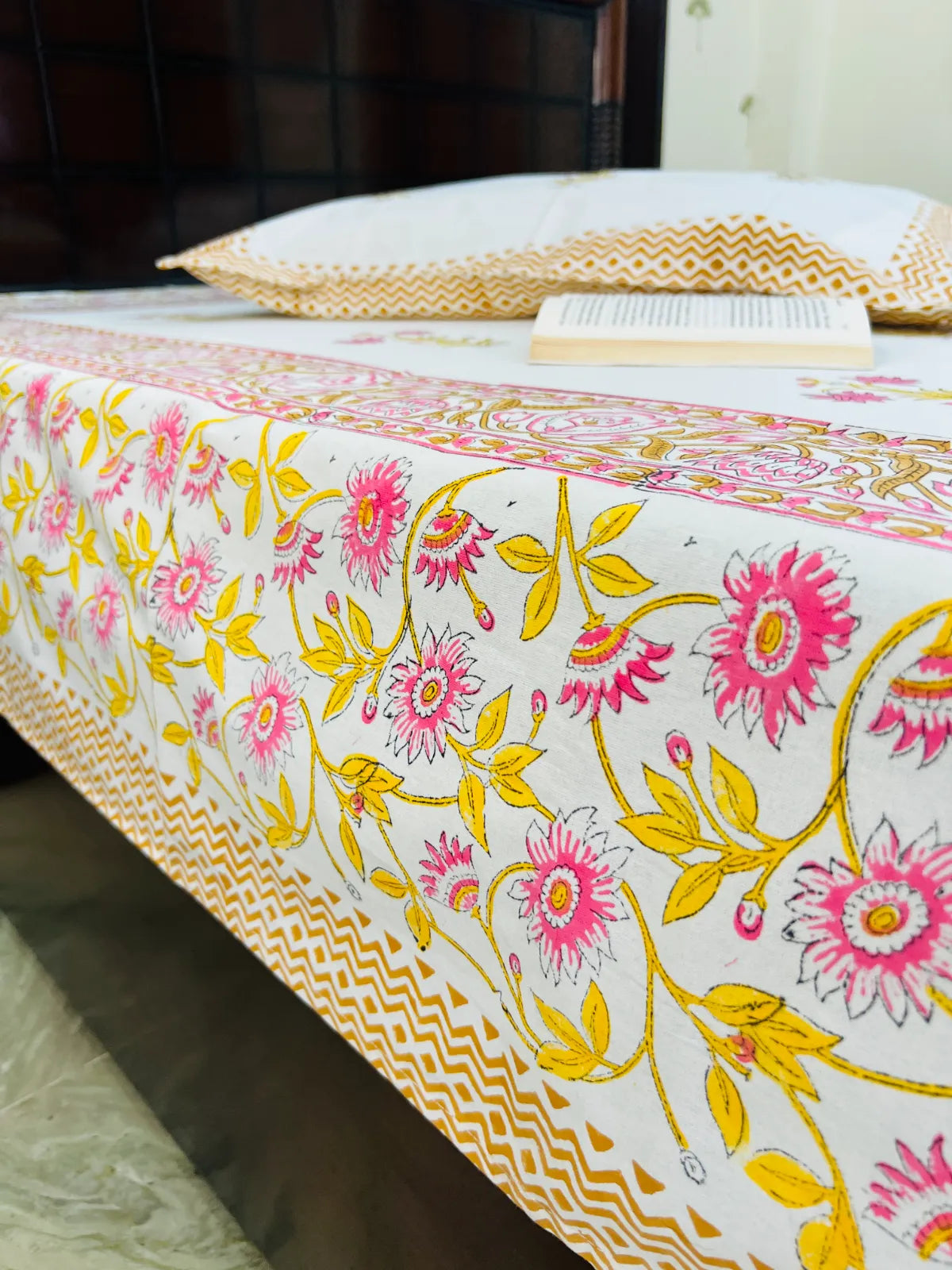 A close-up of a white bedspread or tablecloth featuring a vibrant pink and yellow floral pattern, with a matching zigzag border. A white pillow and an open book are visible on the bed in the background.