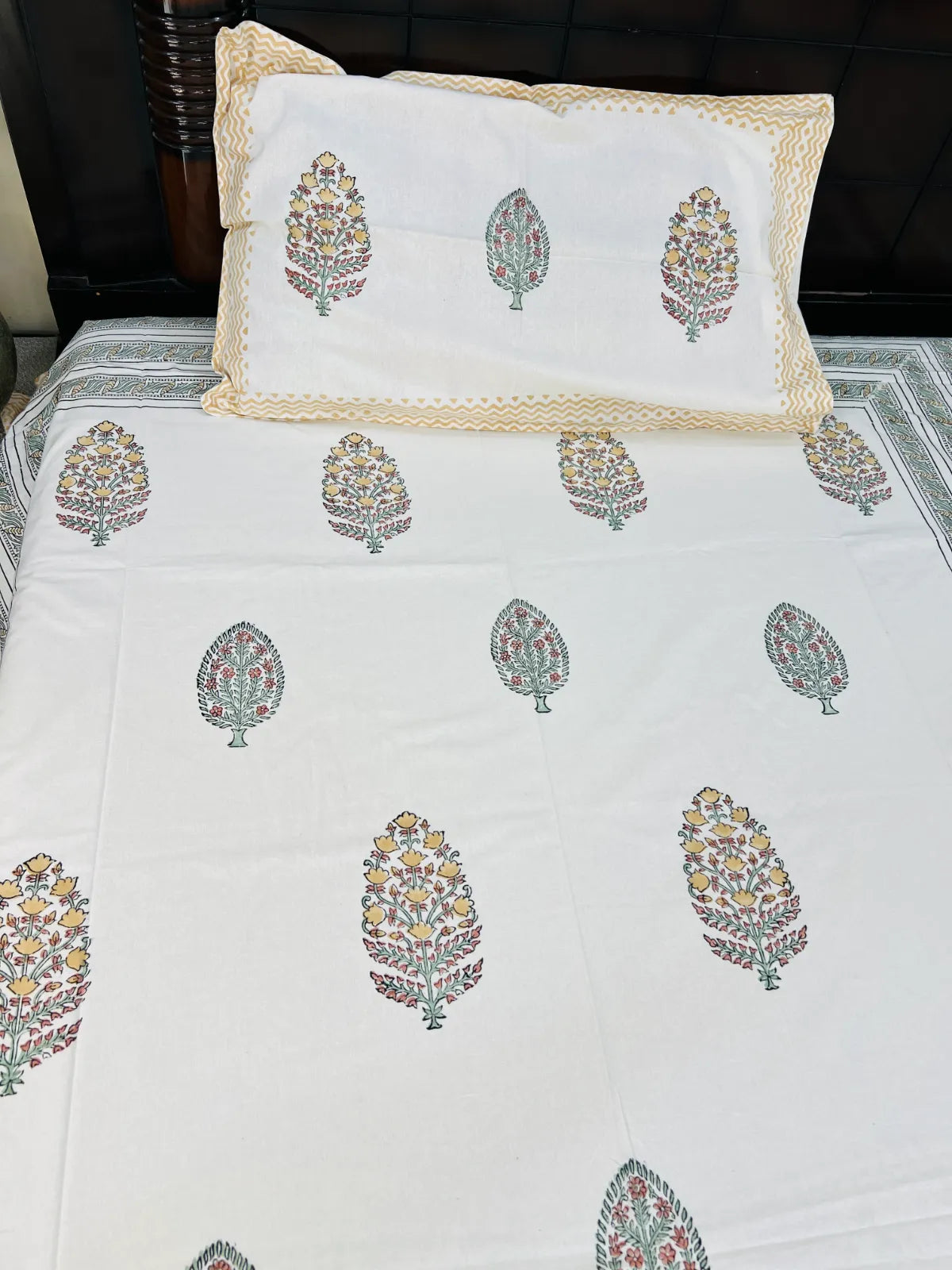 White cotton bed sheet and matching pillowcase featuring a block-printed floral leaf pattern in muted green, pink, and yellow, finished with a beige border.
