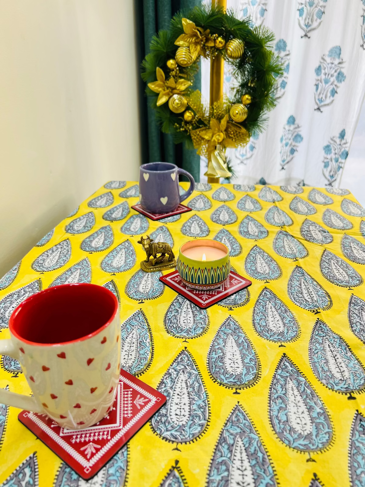 Decorative table setting with mugs, candles, and a wreath on a patterned tablecloth.