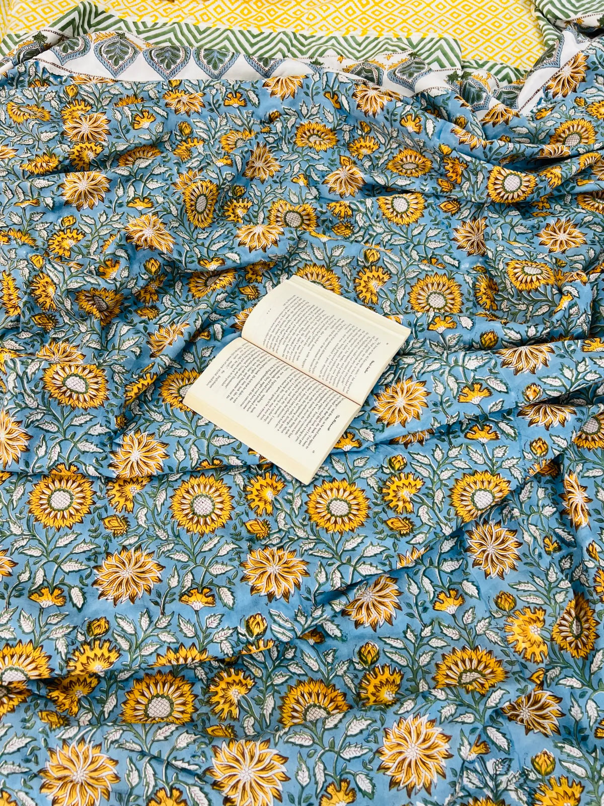 A light blue fabric, likely a quilt or blanket, featuring a dense yellow and white floral block print pattern, styled with an open book on top and a matching patterned textile edge visible at the top.