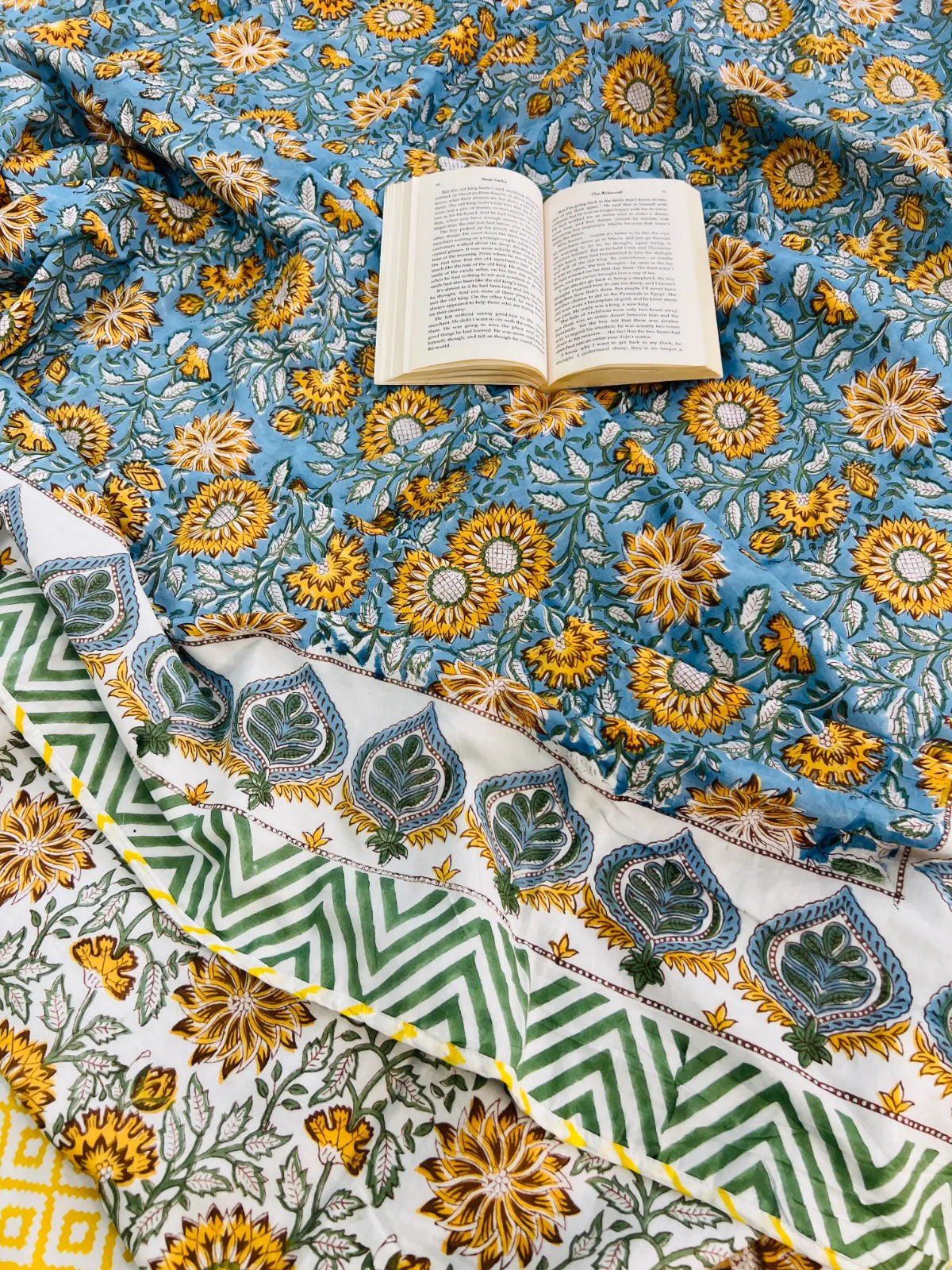 A close-up of a printed fabric with a blue ground, yellow floral motifs, and a decorative border in green, white, and blue. An open book lies on top of the fabric.