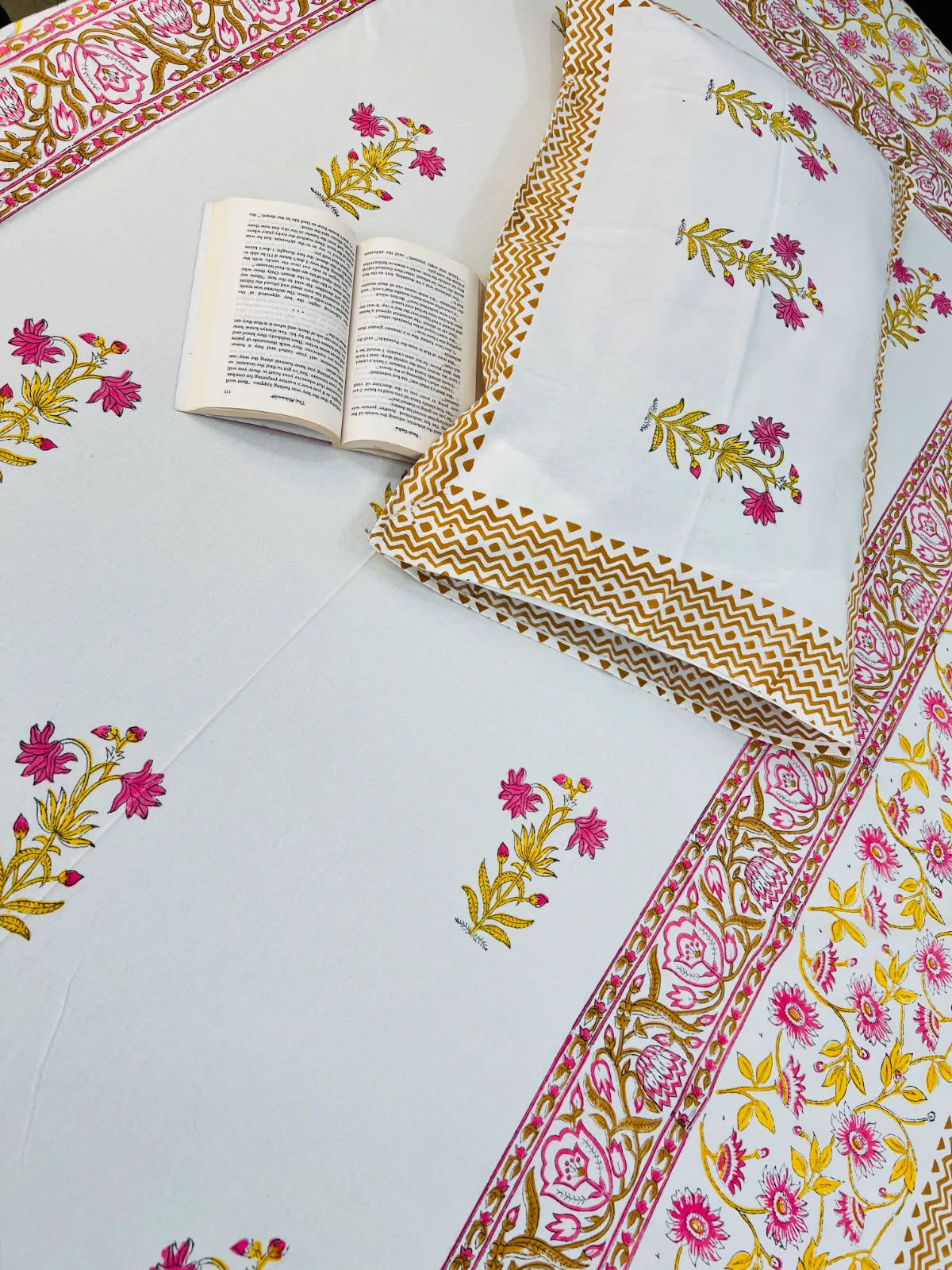A white cotton bedspread or sheet set with pink and yellow floral block prints and a decorative border, shown alongside a matching pillowcase. An open book is placed on the sheet as a styling prop.