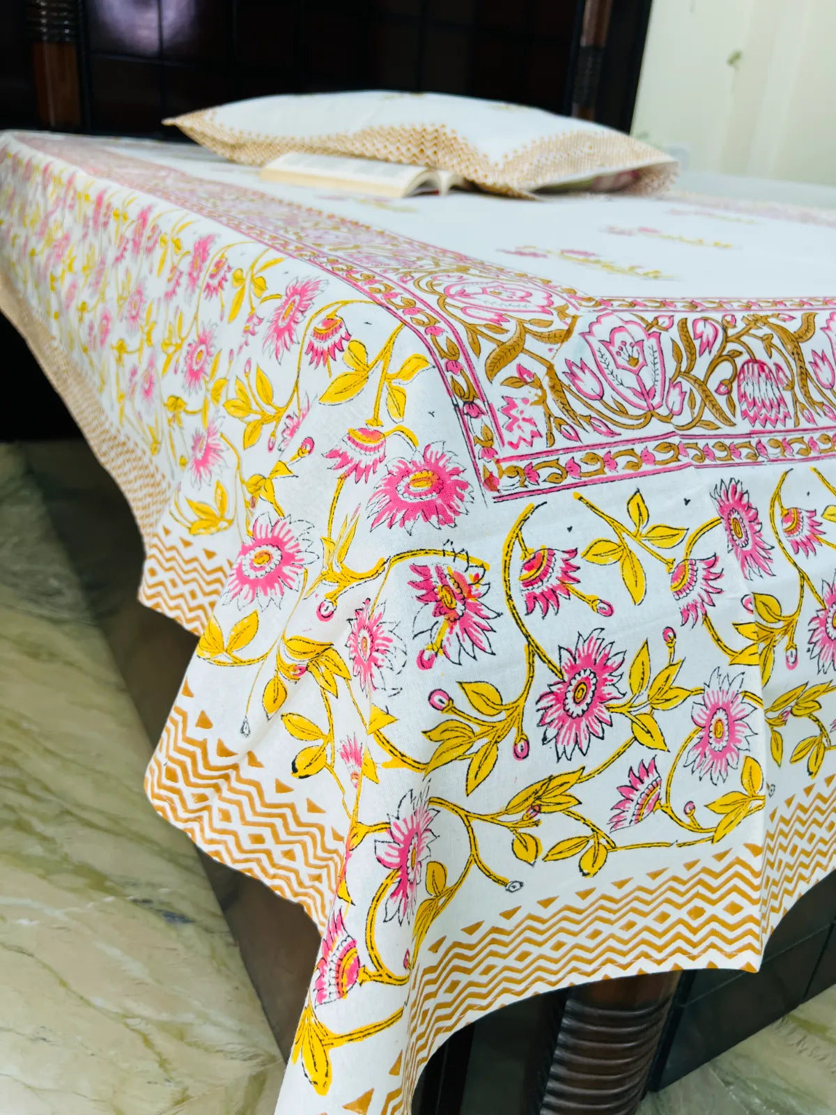 A white cotton tablecloth with a vibrant pink and yellow floral block print pattern, draped over a bed with matching pillows in a bedroom setting.