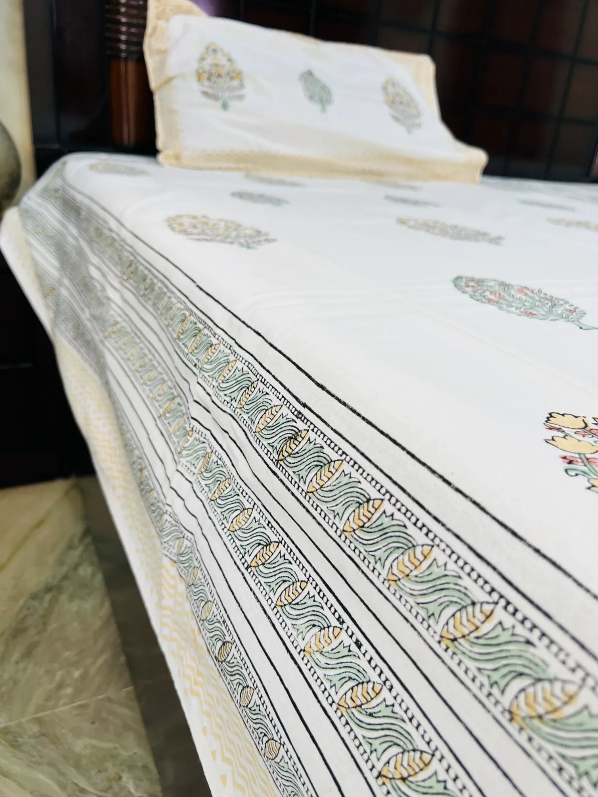 A close-up of a white bed sheet or duvet cover with a decorative border pattern in black, green, and beige, draped over a bed with a matching pillowcase visible at the top.