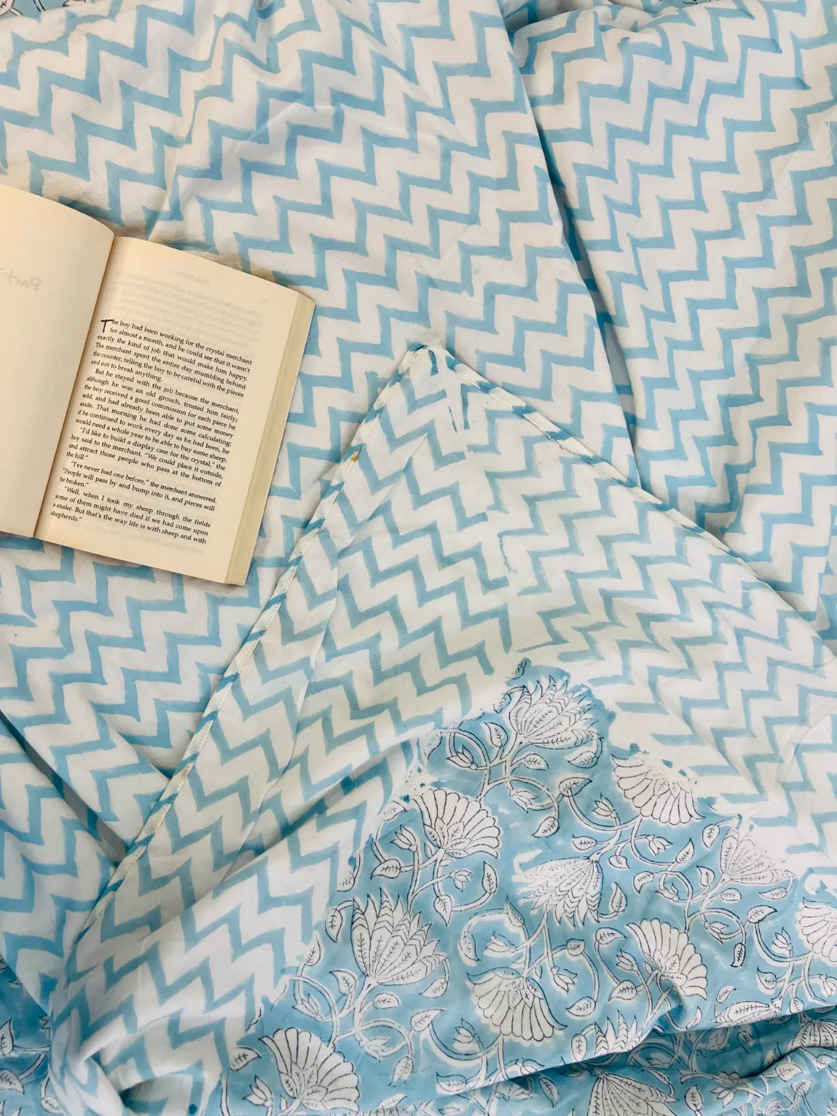 A light blue and white patterned quilt or comforter with a chevron design and a matching pillow featuring a white seashell floral pattern, styled with an open book on top.