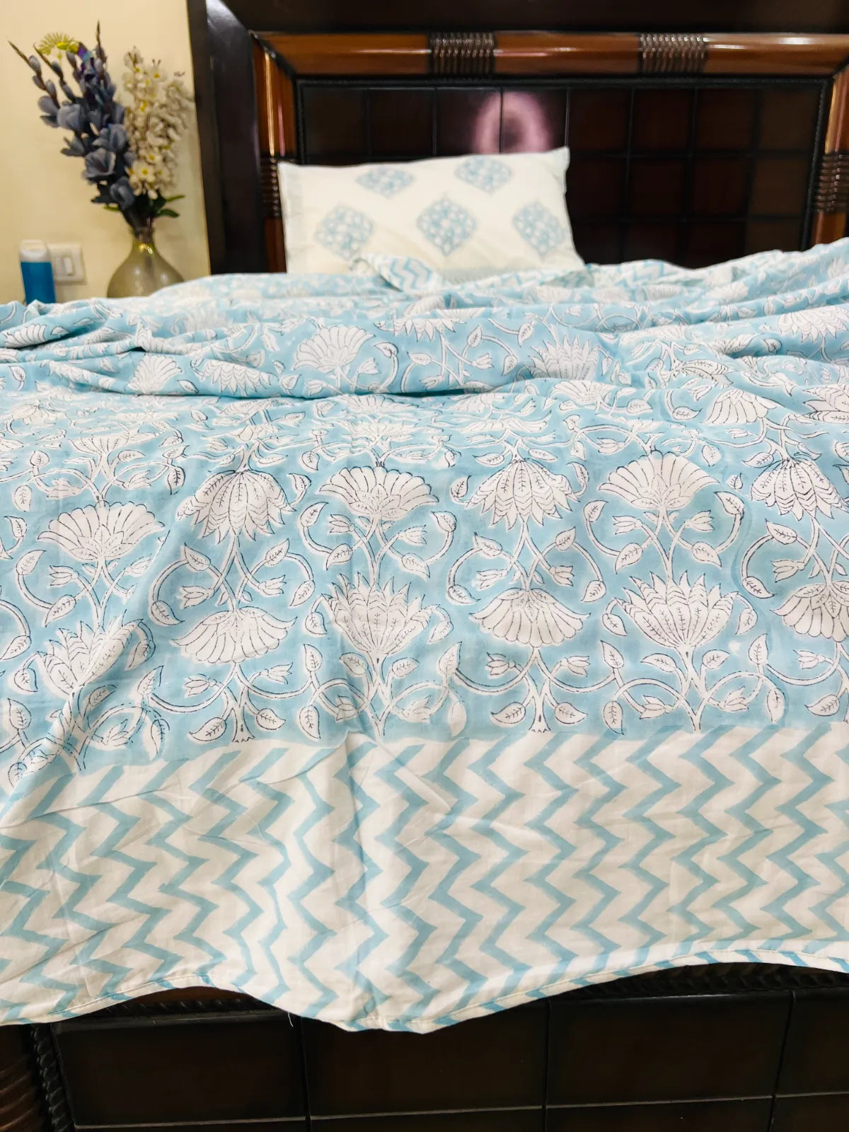 A light blue and white patterned bedspread with a floral motif and a chevron border, shown on a bed with a dark wooden headboard and a matching pillow.
