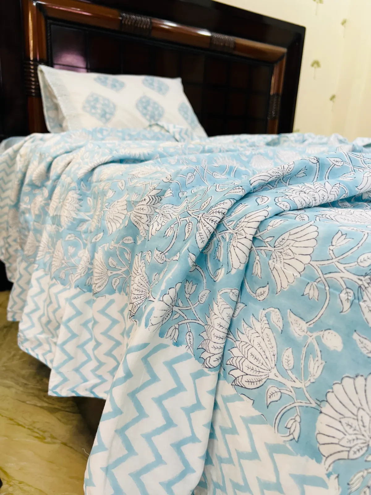 A light blue and white patterned bedspread with a matching pillow, draped over a dark wooden headboard in a bedroom setting.