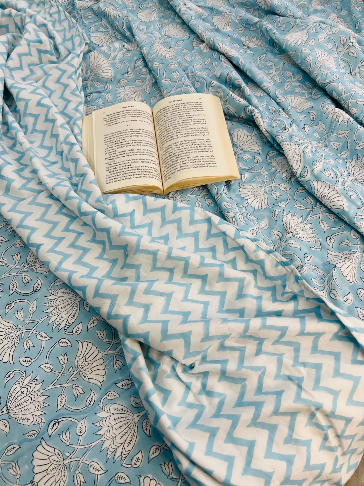 A light blue and white patterned fabric, likely a throw blanket or bedspread, draped with an open book placed on top.