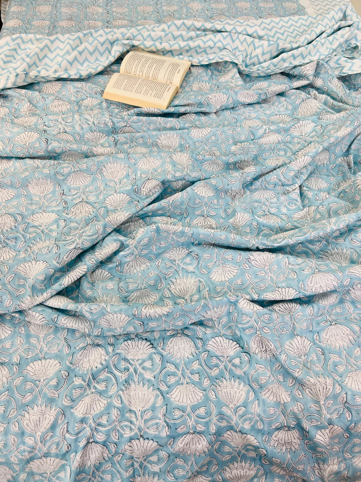A light blue fabric with a white floral block print pattern, draped to show its texture and design. A small open book is placed on top of the fabric.