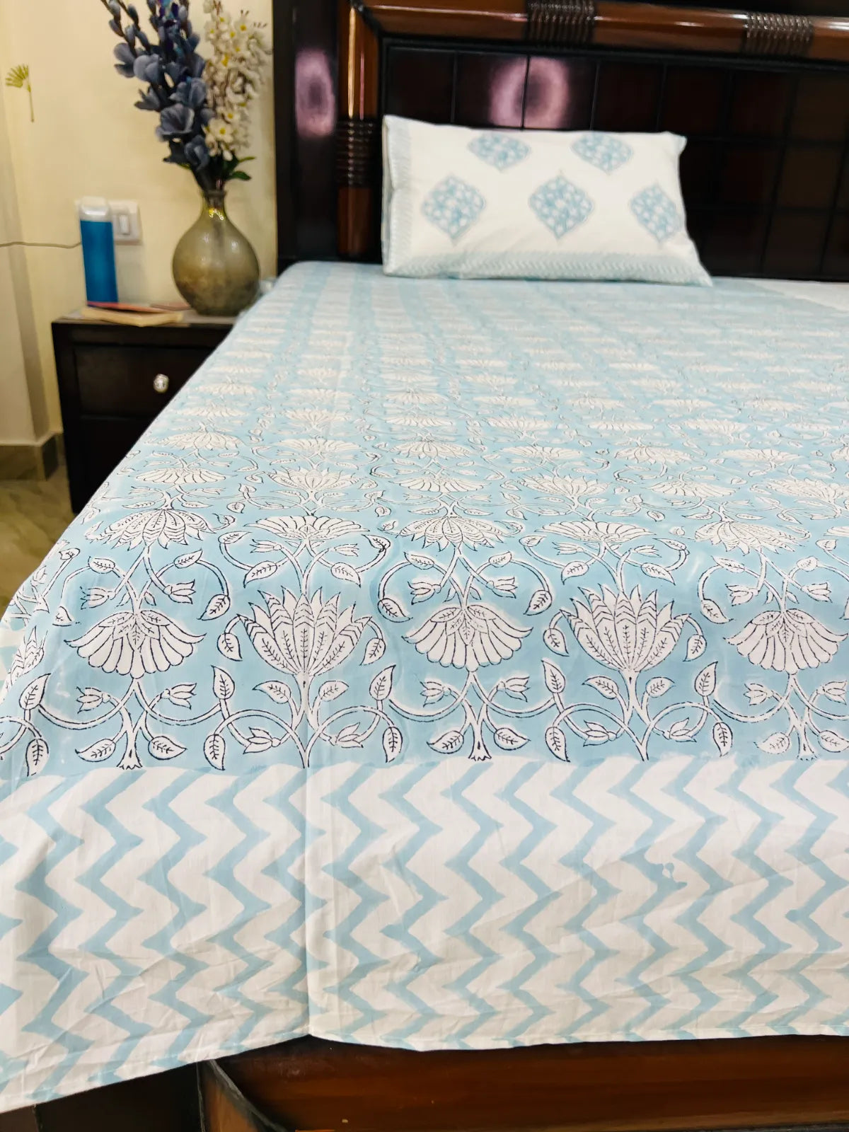 A light blue and white patterned bedspread with matching pillow on a bed with a dark wooden headboard, next to a bedside table with a vase of flowers.