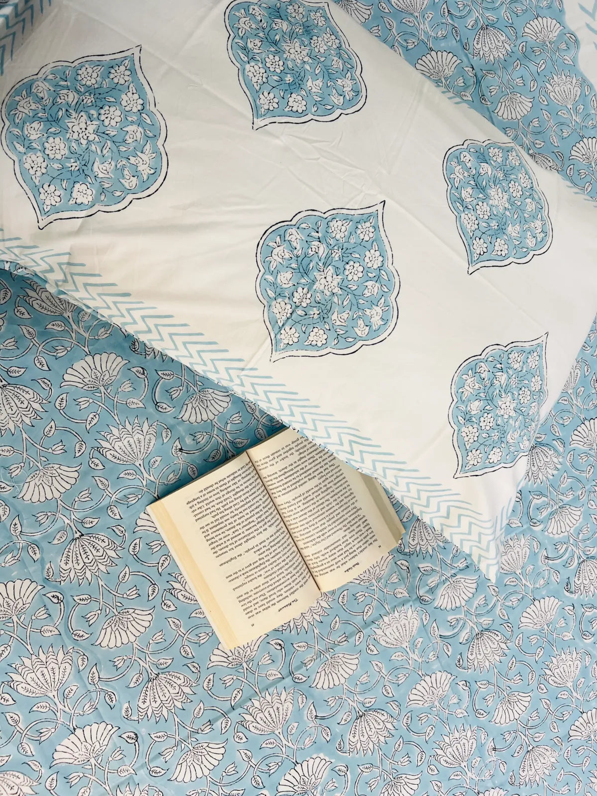 A styled flat-lay of a light blue and white patterned bedding set, featuring a pillow with blue and white floral medallions on a white background, placed on top of a matching patterned quilt. An open book lies on the quilt.