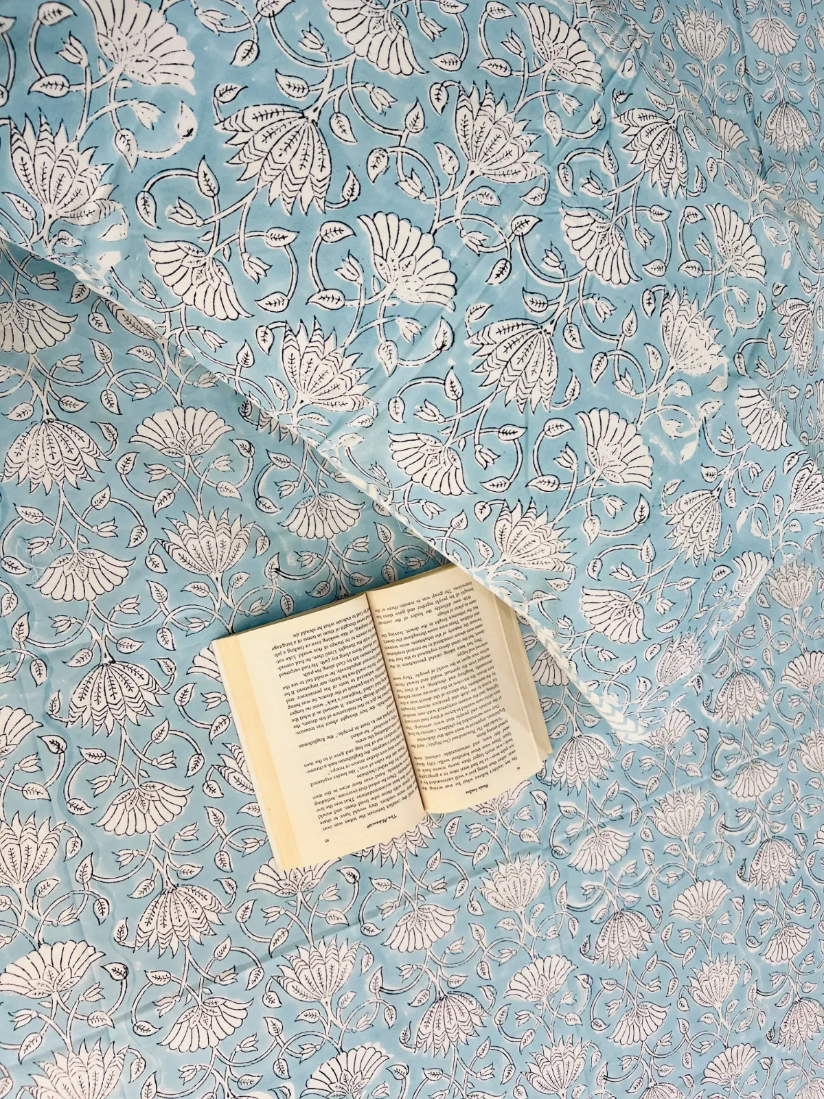 A light blue quilted blanket with a white floral block print pattern, styled with an open book on top.