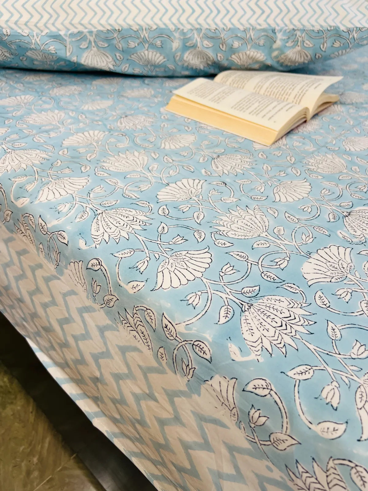 A light blue and white patterned bedsheet with a floral block print design and a matching chevron-patterned border, shown on a bed with an open book on top.