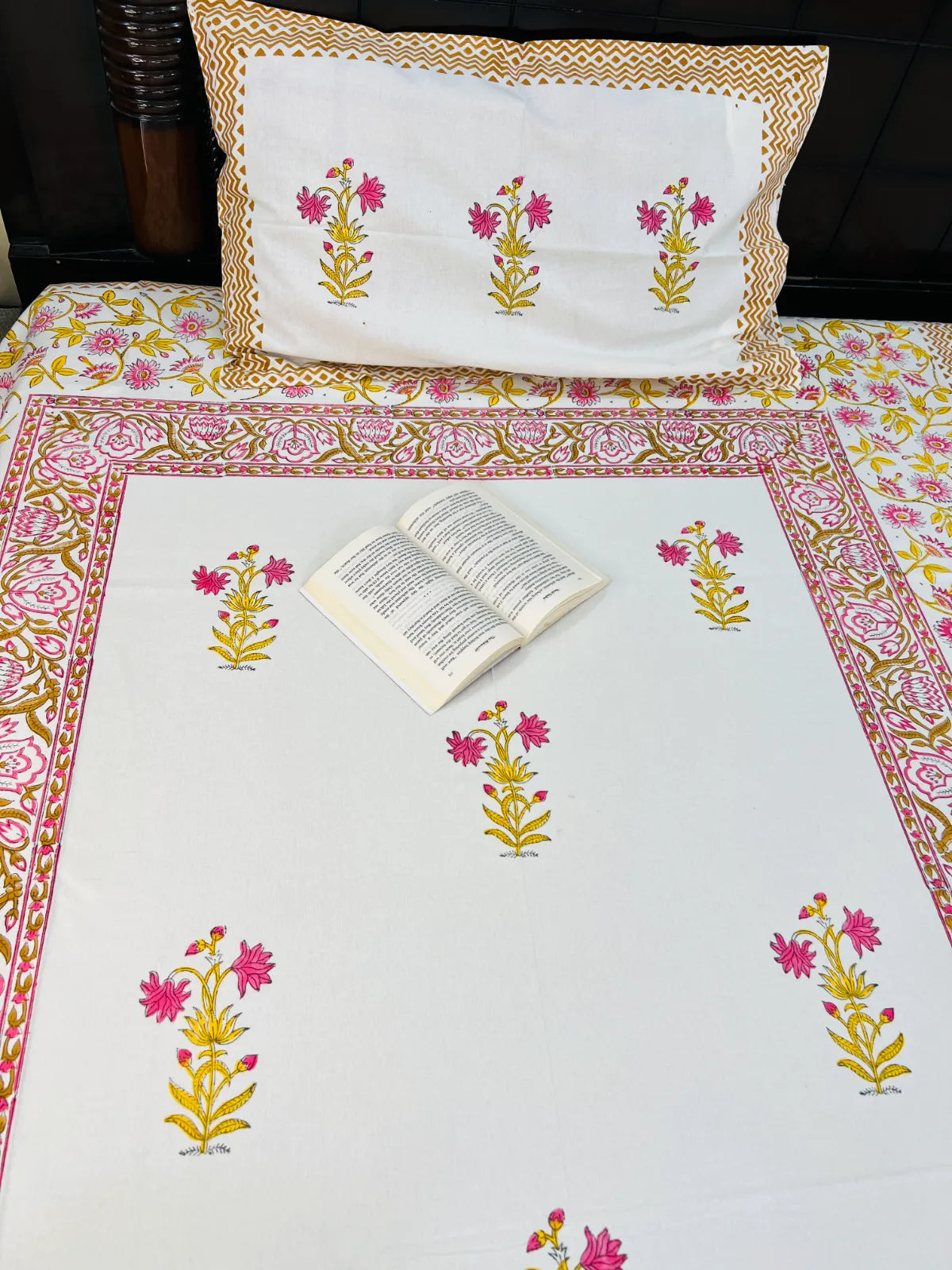 A white cotton tablecloth with a pink and gold floral border and matching floral motifs, styled with a matching decorative pillow and an open book on a bed.